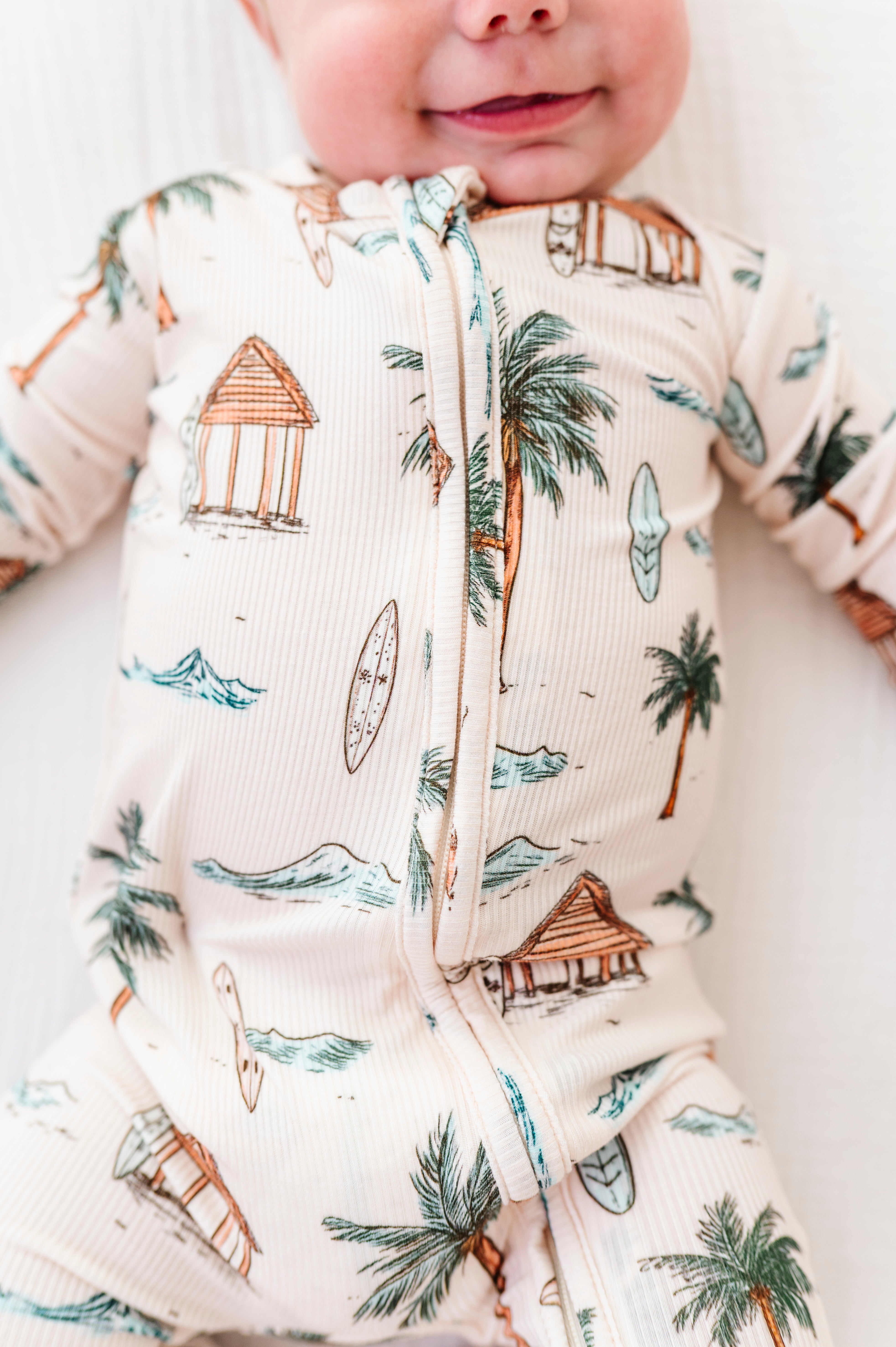 Baby wearing a onesie with beach-themed design, including palm trees and huts.